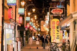 この画像は、夜になって明かりがともった日本の商店街を描いています。色とりどりの看板や提灯が並び、人々がぶらぶらと歩いています。企業が今こそ活用すべき「note」！ブランディングと SEO 対策の重要性と同じように、この商店街も独自の雰囲気や個性を活かして人々を惹きつけています。「note」では、企業が自社のブランド性を伝え、SEO 対策を通じて検索順位を上げることで、より多くの人に知ってもらうことができます。