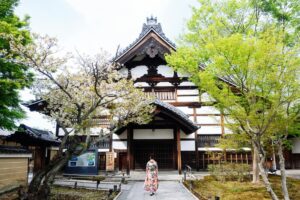 この画像は、古き良き日本の伝統建築が映し出されており、白と茶色の調和が美しい建物の前には、桜の木と新緑の木があり、一人の女性が着物を着て歩いています。このように伝統的な魅力が現代に生き生きと息づいている姿勢と同じように、Facebookマーケティングの再評価！B2B企業が活用すべき3つの戦略は、現代のビジネス環境においても、Facebookの持つ特性を生かして、新たな接客やマーケティングの機会を広げることができます。